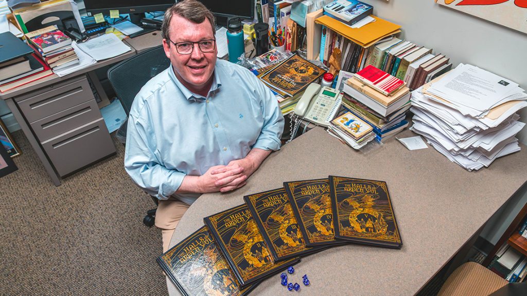 Dr. Rick Barton at his work desk with a set of books and Dungeons and Dragons dice.